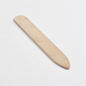Könnte beinhalten: Ein einzelner, glatter, länglicher Kragenstäbchen mit goldfarbenem Metallic-Finish. Das Objekt hat eine abgerundete Oberkante und eine spitz zulaufende Unterkante und bietet ein flaches, schlichtes Design, um die Form eines Hemdkragens zu erhalten.