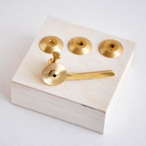 May include: A set of four brass components consisting of three circular washers with central holes and one lever-style handle with a small screw mechanism. The brass pieces have a smooth, metallic finish and are arranged on a light wood block.