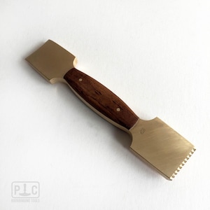 May include: A double-ended bookbinding tool featuring a central dark brown wooden handle secured with two metal pins, flanked by two flat brass heads. One head is smooth and rectangular, while the other features a serrated edge. PLC is stamped on one side.