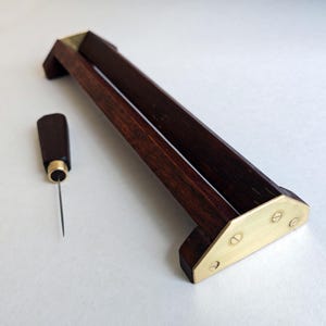 Könnte beinhalten: Ein dunkelbrauner Holzständer mit einem langen horizontalen Schlitz und dreieckigen Messing-Endplatten, die mit Schrauben befestigt sind, sowie ein passendes Werkzeug mit dunkelbraunem Holzgriff und einer dünnen Metallnadel.