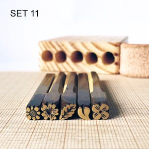 Könnte beinhalten: Ein Set aus fünf Metallstempelwerkzeugen mit der Aufschrift SET 11, die jeweils ein eigenes graviertes Motiv an der Spitze aufweisen, darunter eine Sonnenblume, ein Gänseblümchen, ein Blatt, ein Herz und eine vierblättrige Blume, in einer Reihe.