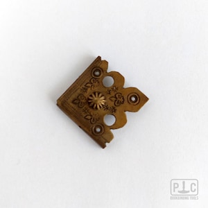 Könnte beinhalten: Ein messingfarbener Bucheckenschutz aus Metall mit geprägtem Blumenmuster, einem zentralen erhabenen runden Knopf und drei kleinen runden Löchern zur Befestigung. Das Teil hat eine rechtwinklige Form, die über die Ecke eines Buches passt.