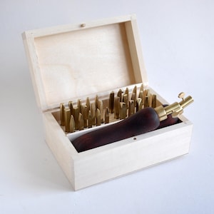 Puede incluir: Una caja de almacenamiento de madera clara que contiene una colección de herramientas de estampado de latón con varias formas de punta y una herramienta con mango de madera marrón oscuro y cabeza de latón con un tornillo de ajuste.