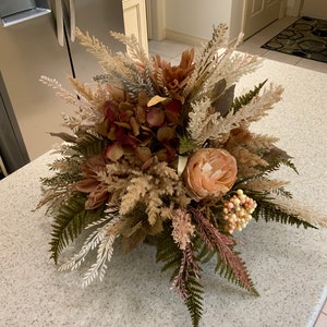 May include: A dried floral arrangement in a brown basket with beige, brown, and pink flowers and greenery.