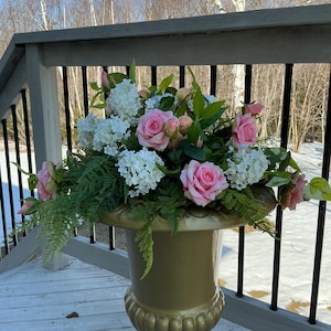 May include: A gold-coloured urn filled with a floral arrangement. The arrangement includes pink roses, white hydrangeas, and green foliage. The urn sits on a wooden deck with a black railing, with trees and a bright sky in the background.