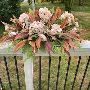 May include: A decorative floral display with light pink hydrangeas, roses, and green foliage. The arrangement incorporates reddish-brown leaves and is placed on a wooden deck, with a natural outdoor background.