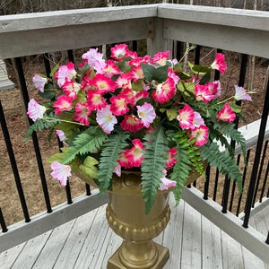 May include: A gold-colored urn-shaped planter filled with vibrant pink and white flowers and green fern-like foliage. The planter sits on a wooden deck with a white and gray painted finish, surrounded by a black railing.