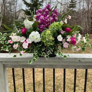 Puede incluir: Un vibrante arreglo floral en una maceta negra, con una mezcla de flores moradas, blancas, rosas y verdes. El arreglo incluye hortensias, guisantes de olor e hiedra, creando una exhibición exuberante y colorida. El arreglo está sobre una barandilla de madera gris.