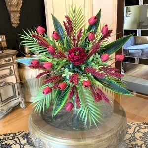 May include: A large, red and green floral arrangement in a clear glass vase. The arrangement features red tulips, green leaves, and red berries. The arrangement is sitting on a round, wooden table with a glass top.