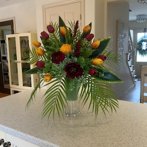 May include: A floral arrangement in a clear glass vase with red, orange, and yellow tulips, burgundy roses, and green foliage.