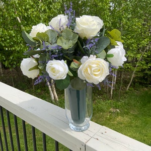 May include: A bouquet of white roses and purple flowers arranged in a clear glass vase. The vase is sitting on a white wooden railing.