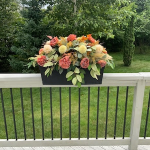 May include: A black planter box filled with a floral arrangement of peach, cream, and brown flowers. The planter is sitting on a white wooden railing.