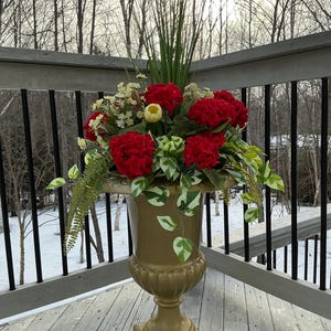 May include: A gold urn-shaped planter filled with bright red flowers, yellow accents, and green foliage. The planter has a pedestal base and is decorated with leaf patterns. The arrangement sits on a wooden deck with a winter backdrop.