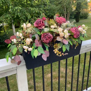 May include: A black rectangular planter box overflowing with faux flowers and greenery. The arrangement includes large burgundy peonies, cream-colored roses, and various green and burgundy leaves. The planter sits on a white railing with black vertical bars.