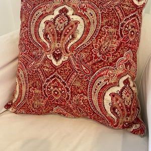 May include: A square decorative pillow with a red, beige, and burgundy paisley pattern. The pillow features a detailed, ornate design with floral and teardrop shapes. The pillow is resting on a white surface.