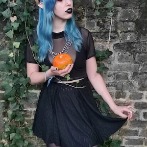 May include: A woman with blue hair and elf ears is wearing a black mesh top with a silver chain necklace and a black skirt. She is holding a small orange pumpkin in her hand.