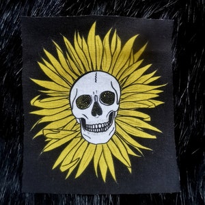 Puede incluir: Un parche de tela negro con una calavera blanca dentro de un diseño de girasol amarillo.