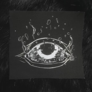 May include: A black patch with a white eye graphic. The eye is surrounded by white lines that resemble water droplets.