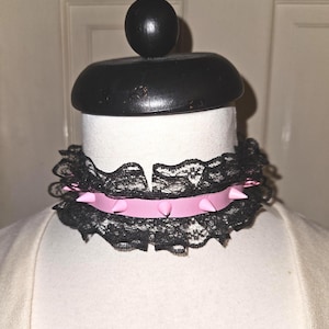 May include: A black lace choker with a pink band and pink spikes. The choker is displayed on a white mannequin head with a black wooden top.