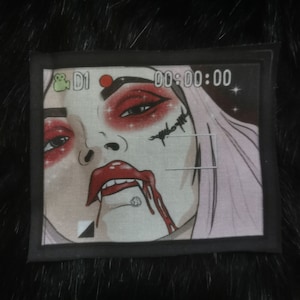 May include: A black and white embroidered patch featuring a close-up image of a woman's face. The woman has red lipstick, red eyeshadow, and a bloody lip. The image is framed in a black rectangle with a camera icon and the text "D1 00:00:00".