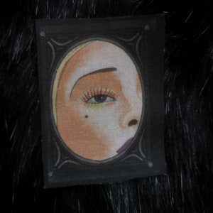 Könnte beinhalten: Ein kleiner, rechteckiger Aufnäher mit schwarzem Rahmen, der eine stilisierte Illustration eines Frauengesichts zeigt. Das Bild zeigt ein Frauenauge mit dunkler Wimpernlinie und gelbem Lidschatten. Der Aufnäher hat dekorative Elemente.