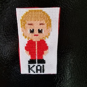May include: Cross-stitch portrait of a person with blonde hair and a red jacket. The name "KAI" is stitched in black block letters at the bottom. The background is white with a red border.
