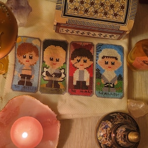 May include: A white canvas with five embroidered squares featuring cartoon characters. Each square has a different character with a unique design and color scheme. The characters are all male and have large eyes and smiles. The canvas is surrounded by various objects, including a gold decorative bowl, a wooden box, and a clear glass bottle.