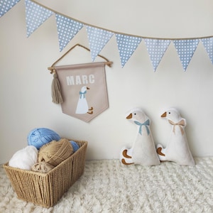 Op de afbeelding: Een beige stoffen wandbanner met de naam MARC en een ganzenillustratie, twee ganzenvormige pluche kussens, een gevlochten mand gevuld met bollen wol en een blauw-witte geblokte vlaggenlijn die boven de items hangt.