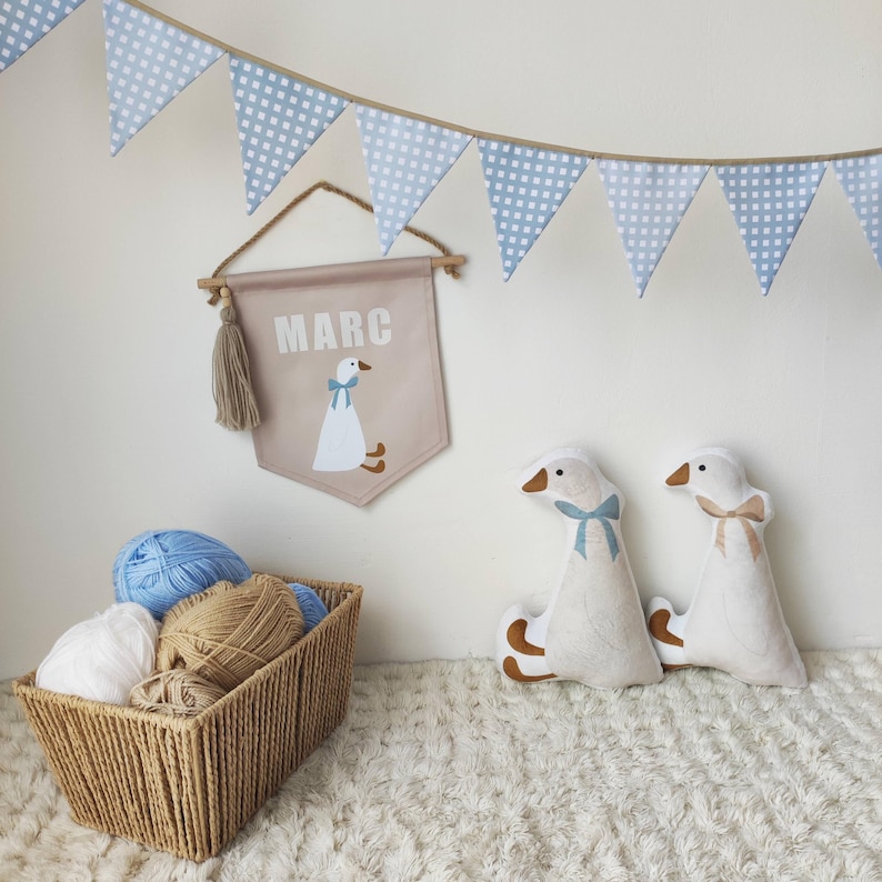 Op de afbeelding: Een beige stoffen wandbanner met de naam MARC en een ganzenillustratie, twee ganzenvormige pluche kussens, een gevlochten mand gevuld met bollen wol en een blauw-witte geblokte vlaggenlijn die boven de items hangt.