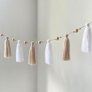 May include: A decorative garland featuring alternating white and beige yarn tassels suspended on a thin cord. The design includes natural light-coloured wooden beads of varying sizes interspersed between the tassels along the length of the string.