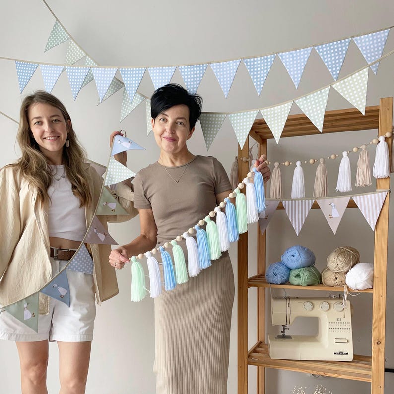 Op de afbeelding: Een decoratieve vlaggenlijn met lichtblauwe en groene stoffen driehoeken met stippen, samen met een slinger van houten kralen en afwisselende witte, lichtblauwe en mintgroene garenkwastjes, inclusief extra stoffen wimpels en garenkwastjes.