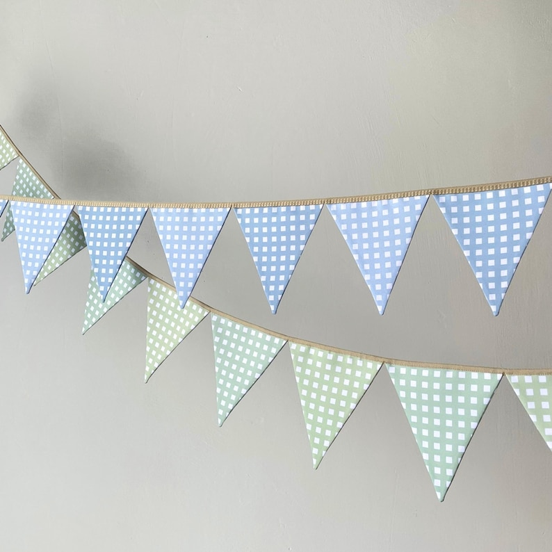 Op de afbeelding: Een decoratieve vlaggenlijn bestaande uit twee rijen driehoekige vlaggetjes. De bovenste rij bevat lichtblauw met witte ruitjespatronen, terwijl de onderste rij lichtgroen met witte ruitjespatronen heeft, allemaal bevestigd aan een beige koord.