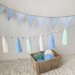Op de afbeelding: Een decoratieve slinger met lichtblauw-witte ruitjesvlaggetjes, opgehangen boven een koord met houten kralen en garenkwastjes in wit, lichtblauw en mintgroen. Een gevlochten mand bevat bollen garen in bijpassende kleuren.