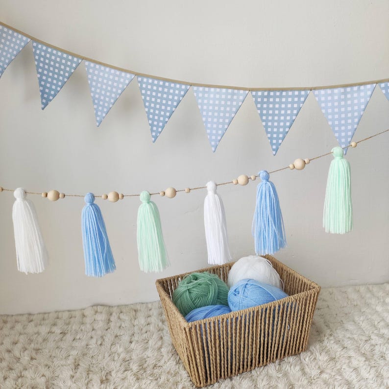 Op de afbeelding: Een decoratieve slinger met lichtblauw-witte ruitjesvlaggetjes, opgehangen boven een koord met houten kralen en garenkwastjes in wit, lichtblauw en mintgroen. Een gevlochten mand bevat bollen garen in bijpassende kleuren.