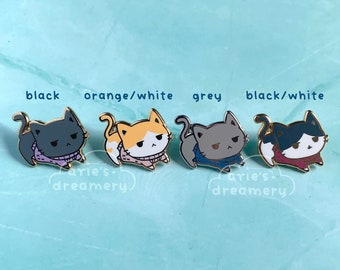 May include: Four enamel pins featuring a cartoon cat wearing a sweater. The cat is shown in four different color variations: black, orange and white, grey, and black and white. The pins are all the same design, but with different color combinations. The pins are on a light blue background.