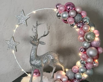 May include: A silver glitter reindeer figurine stands in front of a white, glittery, crescent moon-shaped wreath. The wreath is decorated with pink and silver ornaments and a string of white lights. The base of the wreath is covered in white cotton.