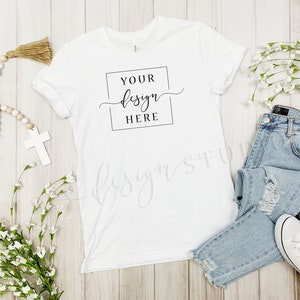 Op de afbeelding: Wit T-shirt met een zwart vierkant en de tekst "YOUR design HERE" op een witte houten achtergrond. Het T-shirt ligt plat met een lichtblauwe jeans, witte sneakers en witte bloemen.