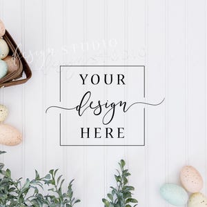May include: A white wood background with a square frame that says "Your design here".  There are green sprigs of greenery on the bottom left and right corners of the image.  There are pastel colored Easter eggs in a brown basket on the top left corner of the image and more pastel colored Easter eggs on the bottom right corner of the image.