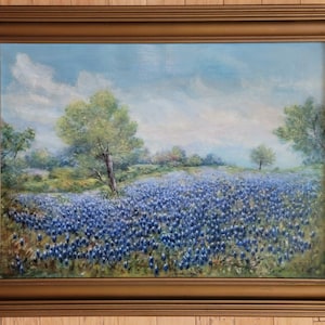 May include: A painting depicting a field of bluebonnet flowers in full bloom. The flowers are in the foreground, with a green hill and trees in the background. The sky is blue with white clouds.