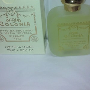 May include: A clear glass bottle of Acqua di Colonia cologne by Officina Profumo S. Maria Novella Firenze 1612. The bottle is filled with a light yellow liquid and has a gold cap. The label is gold with black text and a crest.