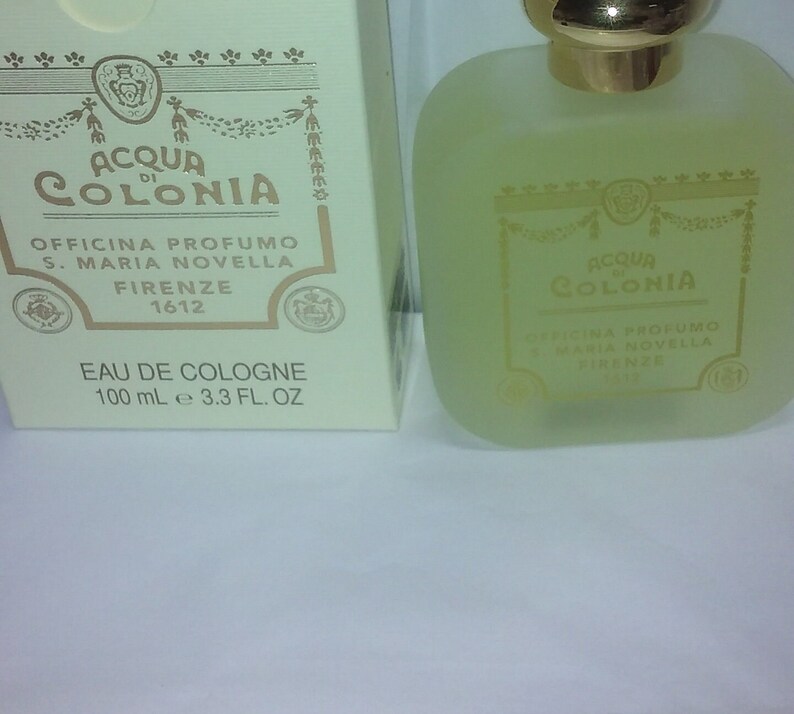 May include: A clear glass bottle of Acqua di Colonia cologne by Officina Profumo S. Maria Novella Firenze 1612. The bottle is filled with a light yellow liquid and has a gold cap. The label is gold with black text and a crest.