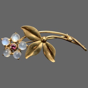 Può includere: Un fermaglio a forma di fiore in oro con una pietra centrale rosa e accenti di pietra di luna bianca.
