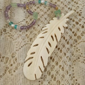 May include: A white carved bone feather pendant with a purple and green beaded necklace. The necklace has gold accents and is on a white lace background.