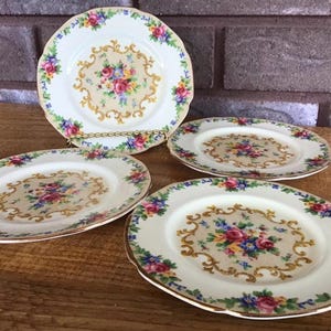 May include: A set of four round ceramic plates featuring a cream-coloured centre with a central floral bouquet of pink, red, and blue flowers surrounded by gold scrollwork. The scalloped rims are decorated with a colourful floral garland pattern.