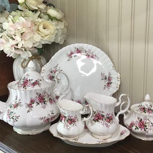 May include: A white porcelain tea set with a floral rose pattern. The set includes a teapot, sugar bowl, creamer, serving plate, and two small cream pitchers. Gold trim accents the edges of each piece.
