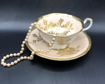 May include: A pale pink porcelain teacup and saucer set featuring gold floral patterns and gilded scalloped edges. A single strand of cream-colored pearls is draped over the rim of the teacup and rests on the saucer, encircling the base.