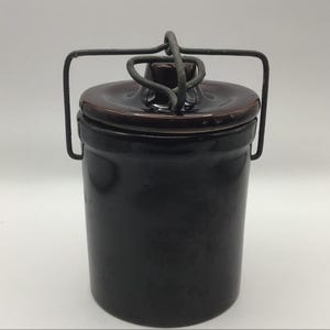 May include: A dark brown ceramic crock with a matching lid and a metal handle. The crock is cylindrical with a slightly tapered shape. The lid has a small knob and the metal handle is attached to the sides.