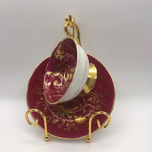 May include: A vintage teacup and saucer set displayed on a gold-coloured metal stand. The teacup and saucer are a deep red with gold floral patterns. The teacup has a white interior. The stand has a curved design.