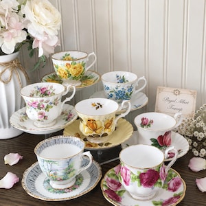 Peut inclure: Une collection de sept tasses à thé et soucoupes Royal Albert avec divers motifs floraux, incluant des roses jaunes, roses et rouges, et des myosotis bleus. Chaque tasse en porcelaine a un bord et une anse dorés, posée sur une surface sombre.