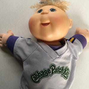 May include: A Cabbage Patch Kids doll with blonde curly hair and blue eyes. The doll is wearing a gray baseball-style jersey with purple and yellow accents and the words "Cabbage Patch Kids" in green.